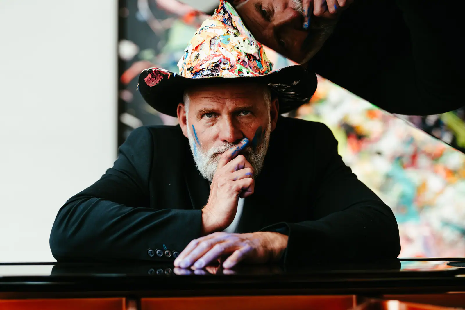 Kunstneren David Bjerre i malingsdækket hat læner sig over et sort flygel foran sine farverige malerier, fotograferet af Skovgaard & Damgaard