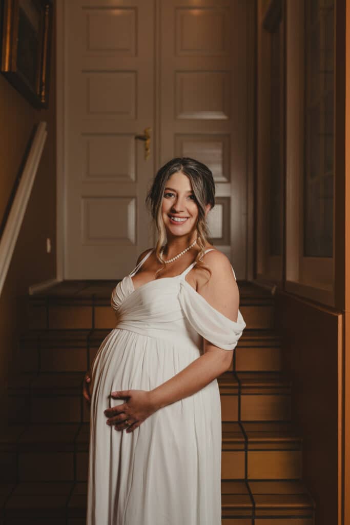 Gravid brud i hvid kjole står på trappe og smiler – bryllupsfotografi af Skovgaard &amp; Damgaard