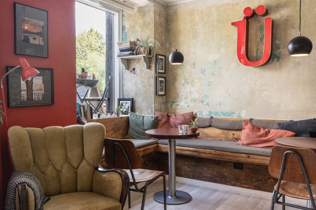 Et hyggeligt siddeområde i en kaffebar med en blanding af vintage møbler og moderne dekorationer. Der er en stor hjørnesofa med farverige puder, et rundt træbord og en vintage lænestol. Et stort vindue lader naturligt lys fylde rummet, mens en rød væg og en stor, rød &quot;Ü&quot;-dekoration pryder væggen.