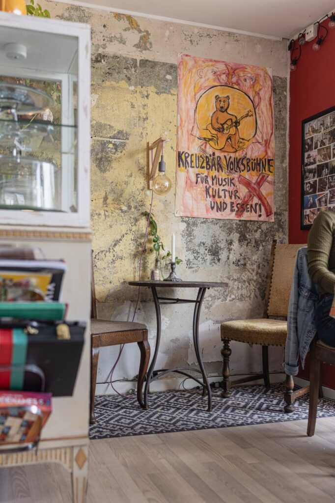 Et hyggeligt hjørne i en kaffebar med en vintage-stil. Et lille bord og to stole står op ad en slidt væg med et håndlavet skilt, der reklamerer for &quot;Kreuzbär Volksbühne&quot; med musik, kultur og mad. I forgrunden ses en del af en montre og nogle bøger.