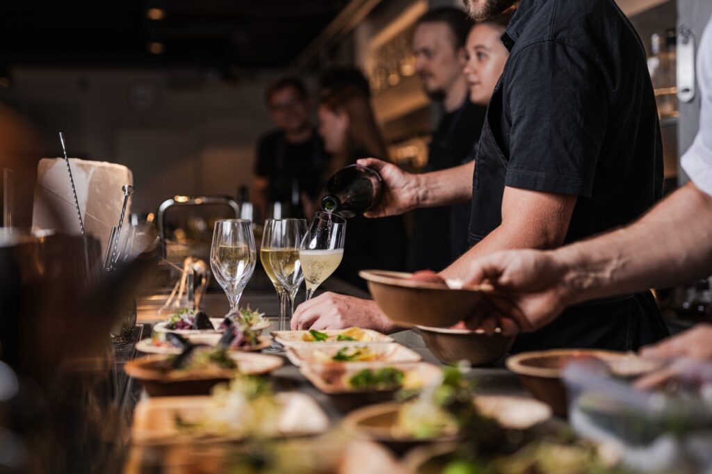 Bag baren på Gedulgt – mad og drikke Bartendere og kokke arbejder sammen bag en bardisk i en cocktailbar. En person hælder vin op i et glas, mens forskellige retter og drinks forberedes på baren foran dem. Der er fokus på både mad og drikke i en stemningsfuld og travl atmosfære.