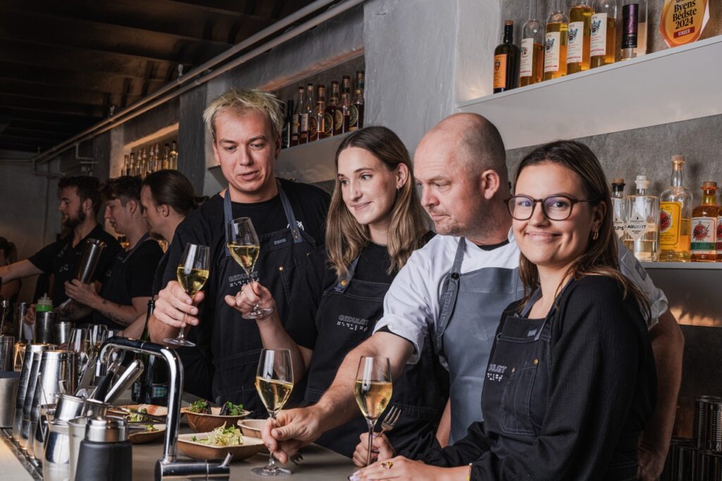 En gruppe bartendere og medarbejdere står bag baren i Gedulgt cocktailbar. De skåler med vinglas, mens de smiler og arbejder sammen. Baren er oplyst med flasker på hylderne i baggrunden, og maden står klar på disken.