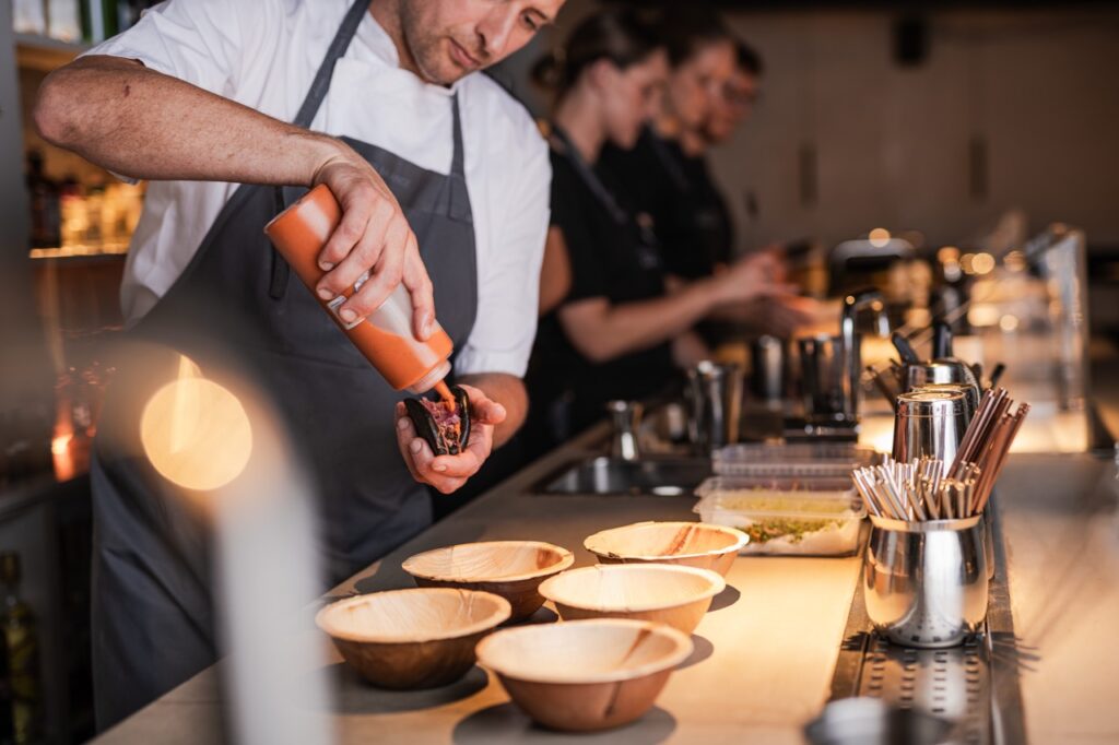 En kok i forgrunden fylder en taco med sauce, mens flere skåle er linet op på bardisken. I baggrunden arbejder andre medarbejdere koncentreret i en cocktailbar.