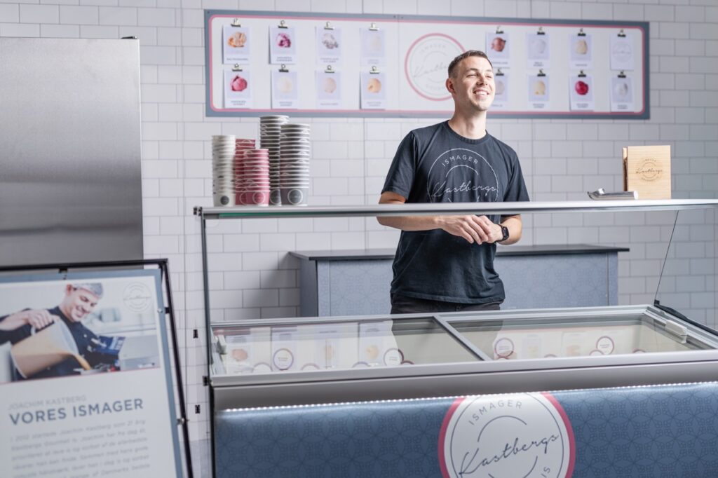 Portrætfotografi af Joachim Kastberg i Kastberg Isbutik Portrætfotografi af Joachim Kastberg bag en isdisk i en moderne isbutik. Han har en t-shirt på med logoet "Kastbergs Gourmet". Ved siden af ham står stablede isbægre, og baggrunden viser en flisebelagt væg med plakater af forskellige isvarianter.