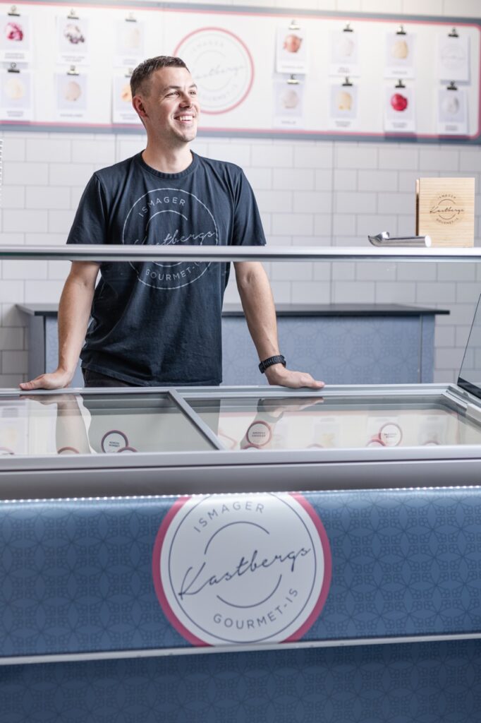 Joachim Kastberg smiler bag en isdisk i en moderne isbutik. Han har en t-shirt p&aring; med logoet "Kastbergs Gourmet" og st&aring;r foran en ren, flisebelagt v&aelig;g med plakater af isvarianter.