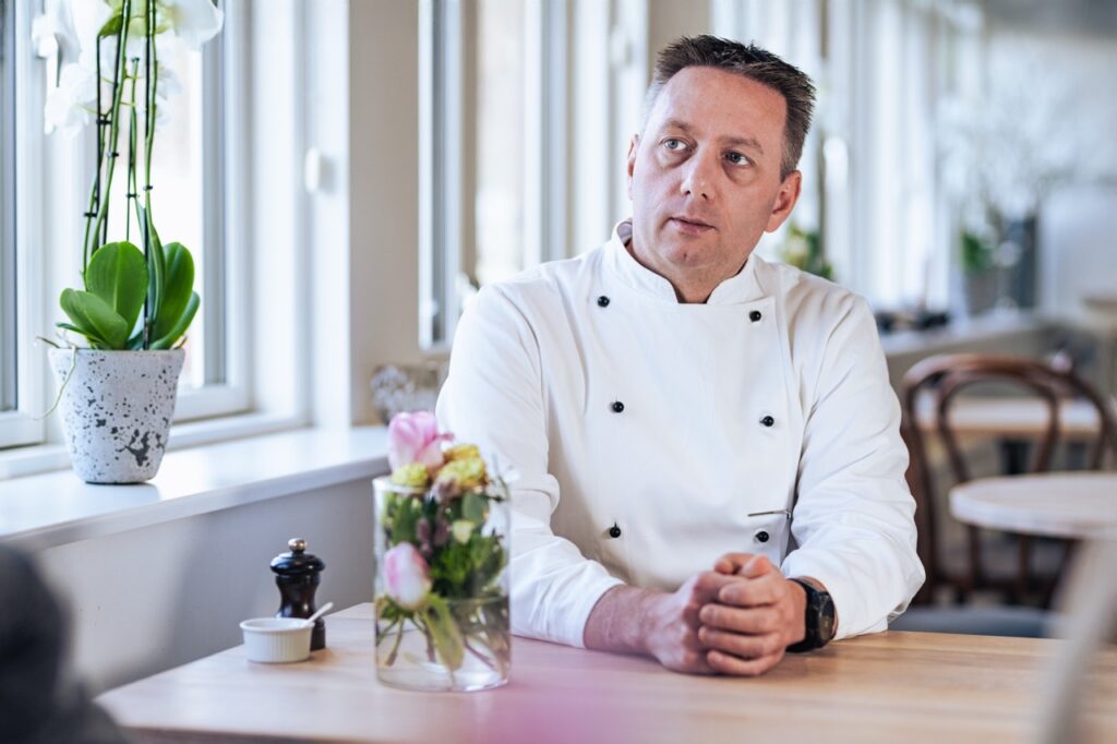 Erhvervsportrætter af Skovgaard & Damgaard – medejer Dean Jespersen fra Knudhule Badehotel i Ry, professionel og indbydende tilstedeværelse Erhvervsportræt af Dean Jespersen, medejer af Knudhule Badehotel i Ry, der sidder ved et bord med blomster, fotograferet af Skovgaard & Damgaard.