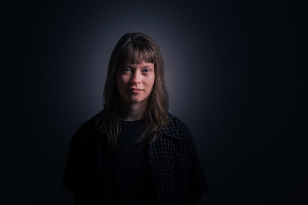 Sangerinde Alex Medhurst portrætteret i studie med mørk baggrund og stemningsfuld belysning, fotograferet af Skovgaard &amp; Damgaard.