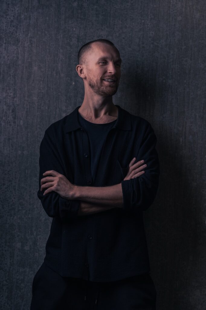 Portrætfotografering af Skovgaard & Damgaard – professionelt erhvervsportræt i mørkt, minimalistisk miljø Stående erhvervsportræt af en person med korslagte arme foran mørk baggrund, fotograferet af Skovgaard & Damgaard.