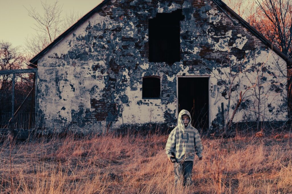 Albumfoto for Drengestreger – forladt hus med barn Albumfoto taget for bandet Drengestreger: et barn i hættetrøje står i højt, tørt græs foran en faldefærdig, hvid bygning med afskallet facade og mørke vinduer – lav sol og kølig farvetone.