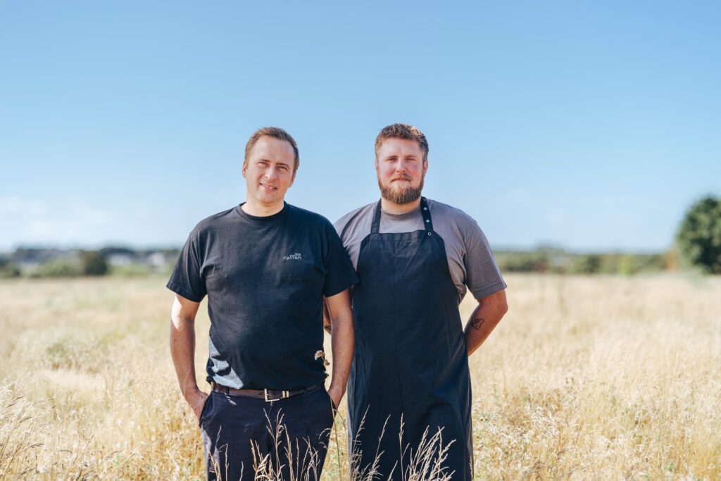 Erhvervsfotografering af Skovgaard & Damgaard – nærbillede af Jacob Møller og Asbjørn Lindhart fra Møller & Damgaard Gastro Jacob Møller og Asbjørn Lindhart fra Møller & Damgaard Gastro står tæt sammen i en gylden mark, fotograferet af Skovgaard & Damgaard.