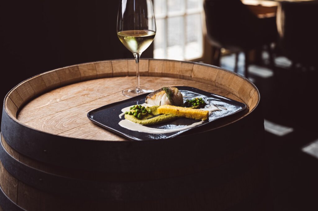 Madfotografi fra Restaurant Coast: tallerken med grønne elementer og fisk – anrettet med hvid sauce og ledsaget af hvidvinsglas, placeret på vintønde i blødt, varmt lys med skygger i baggrunden.