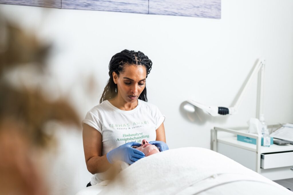 Portrætfoto af Terhas Amare i behandling med klient – iført hvide handsker og t-shirt med logo i klinikrum med behandlingsleje og lampe i baggrunden.