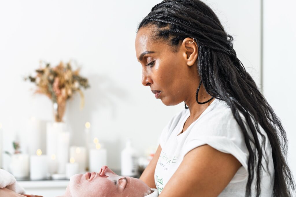 Portrætfoto af kosmetolog Terhas Amare i behandling af klient i klinik – set i profil, med blid koncentration og omgivelser præget af lys, hvide stearinlys og tørrede blomster i baggrunden.