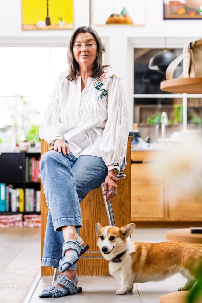 Portrætfoto af foredragsholder Birgitte Damgaard siddende på en høj taburet i et lyst køkkenmiljø, iført hvid bluse og jeans – med en lille hund stående ved fødderne.