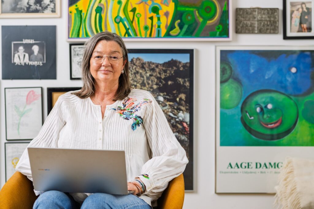 Portrætfoto af Birgitte Damgaard siddende i sennepsgul stol med bærbar computer i skødet – foran væg med kunst og fotos, iført hvid bluse med broderi.