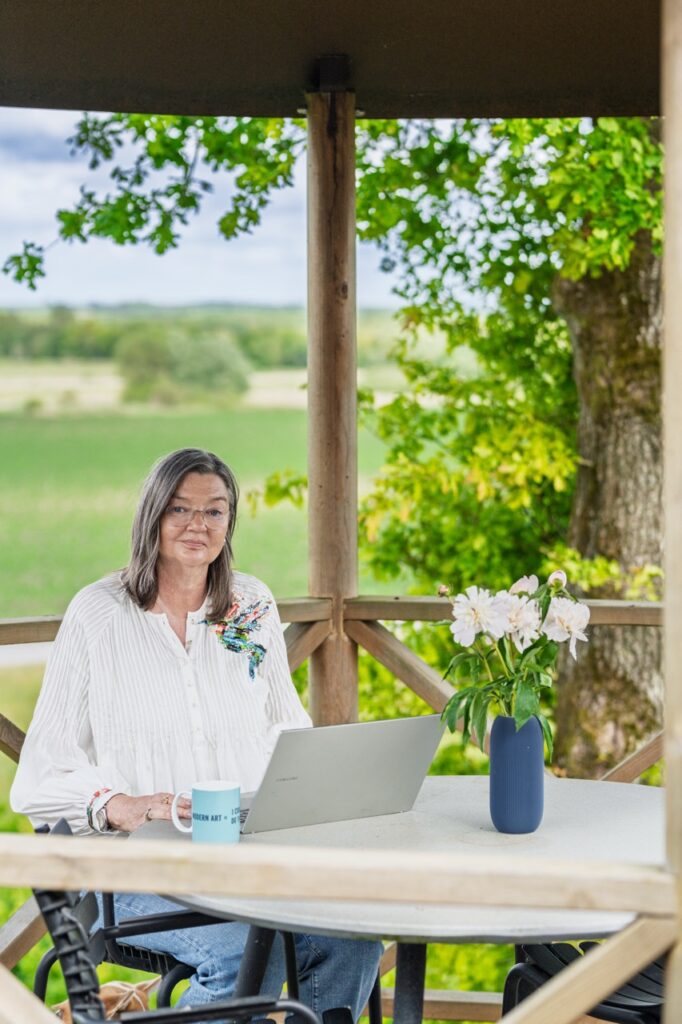 Portrætfoto af Birgitte Damgaard siddende ved rundt bord i udendørs træpavillon med bærbar computer, kaffekop og vase med hvide blomster – med grønne marker i baggrunden.