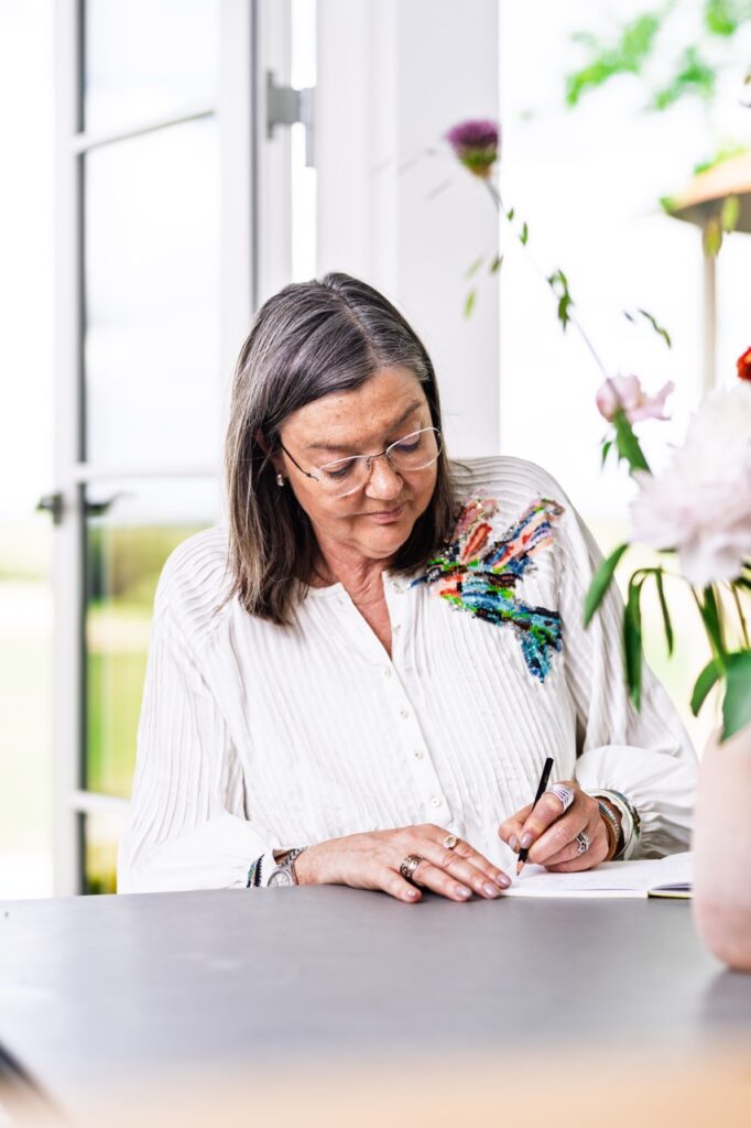 Portrætfoto af Birgitte Damgaard, siddende ved bord og skrivende i notesbog med pen – iført hvid skjorte med broderi, blomster i forgrunden og åbent vindue bagved.