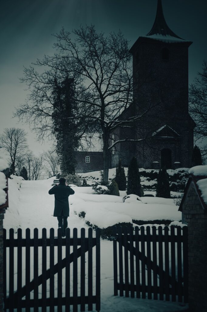 Forsidefoto til Da jeg blinker, er du væk af Jesper Munk Jakobsen Bogforsidefoto til Jesper Munk Jakobsens Da jeg blinker, er du væk: mand i mørk frakke går ad sneklædt sti mod kirke med klokketårn og nøgne træer – fotograferet bag en sort trælåge i vinterligt landskab.