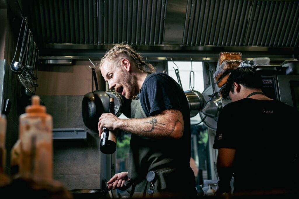 Kokken arbejder i køkkenet på vinbaren Å46 i Aarhus med en flaske vin i hånden, fotograferet af Skovgaard &amp; Damgaard.