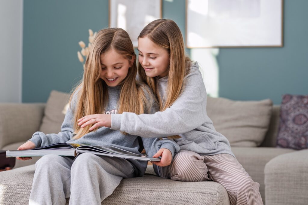 Børneportræt af to piger i gråt tøj, siddende i sofa og bladende i en stor bog sammen – smilende og optagede, i hjemligt miljø med blå væg og dæmpede farver.