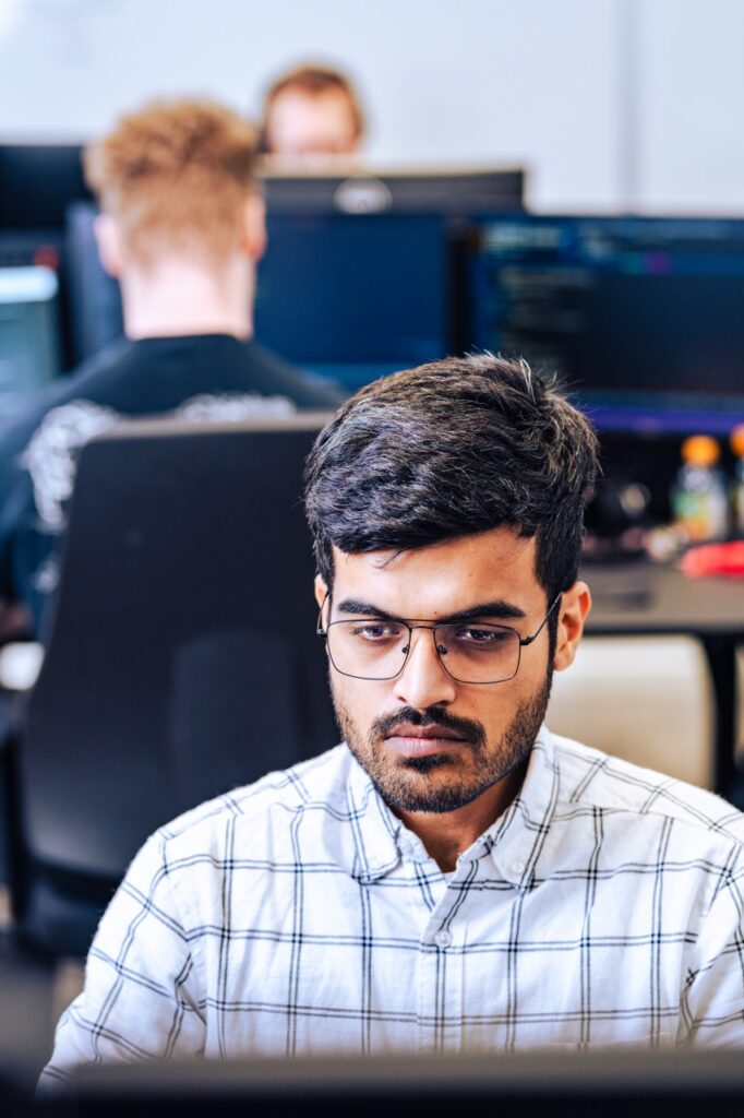 Medarbejder hos UNIwise arbejder koncentreret ved computer med kolleger i baggrunden i kontormilj&oslash;, erhvervsfotograferet af Skovgaard &amp; Damgaard.